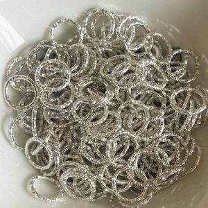 May include: A large pile of silver jump rings, all of the same size, are piled together in a bowl. The rings are textured and have a shiny finish.