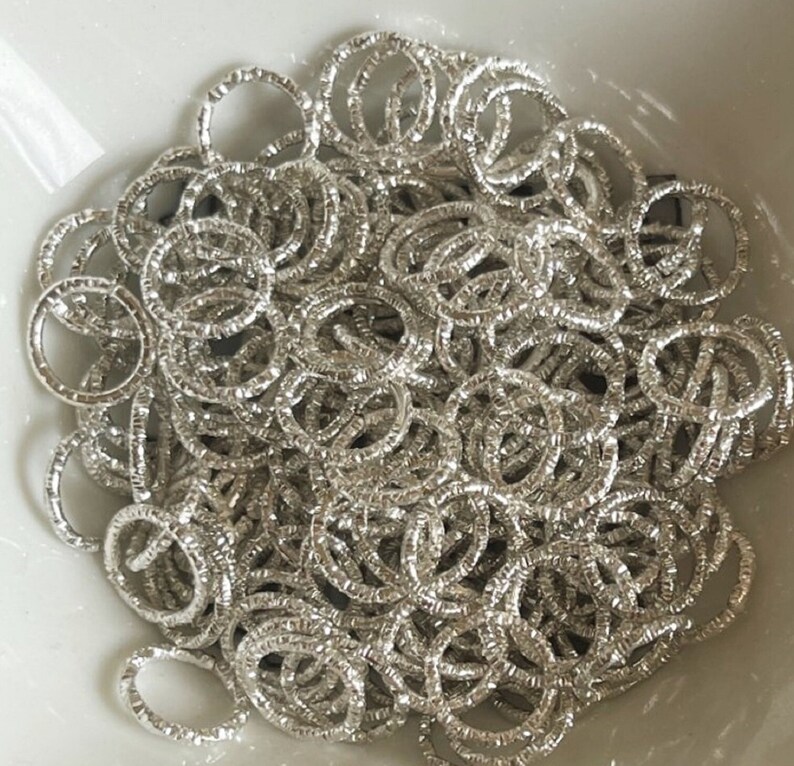 May include: A large pile of silver jump rings, all of the same size, are piled together in a bowl. The rings are textured and have a shiny finish.