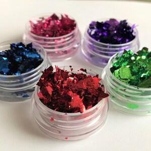 May include: Four small jars filled with colorful metallic flakes. The jars are clear plastic with a silver lid. The flakes are red, blue, green, and purple.
