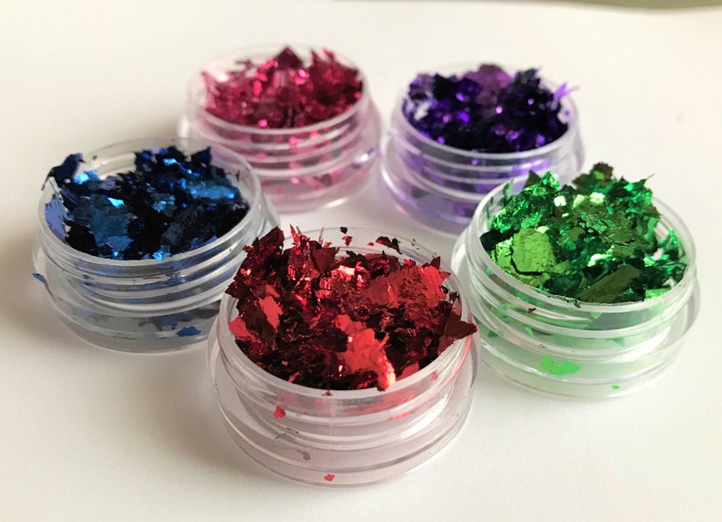 May include: Four small jars filled with colorful metallic flakes. The jars are clear plastic with a silver lid. The flakes are red, blue, green, and purple.