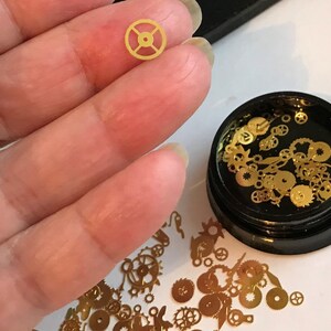 Op de afbeelding: Een klein bakje gevuld met gouden metalen tandwielen en uurwerk onderdelen. Het bakje is zwart met een doorzichtig deksel. Een enkel tandwiel wordt in een hand gehouden ter vergelijking van de grootte.