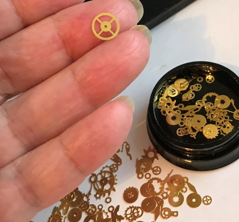 Op de afbeelding: Een klein bakje gevuld met gouden metalen tandwielen en uurwerk onderdelen. Het bakje is zwart met een doorzichtig deksel. Een enkel tandwiel wordt in een hand gehouden ter vergelijking van de grootte.