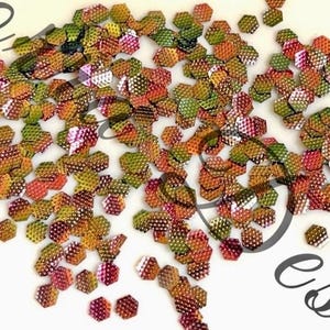 May include: A collection of iridescent, hexagonal sequins in green, orange, and pink hues. The sequins have a textured surface and are scattered on a white background. Suitable for craft projects and embellishments.