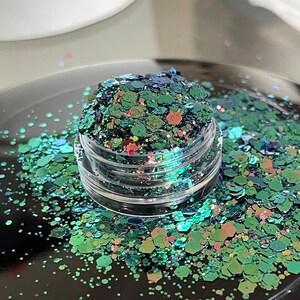 May include: A small, clear jar filled with iridescent glitter. The glitter is a mix of green, blue, and pink hexagonal shapes. Some glitter is scattered around the jar on a black surface. The glitter has a shiny, reflective quality.