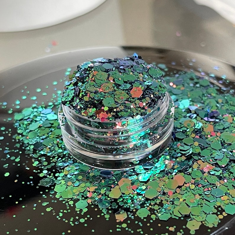 May include: A small, clear jar filled with iridescent glitter. The glitter is a mix of green, blue, and pink hexagonal shapes. Some glitter is scattered around the jar on a black surface. The glitter has a shiny, reflective quality.