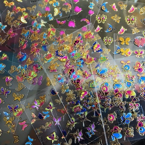 May include: Assortment of colorful butterfly nail stickers on clear plastic sheets. The stickers feature gold accents and various designs, including hearts, flowers, and swirls. The sheets are labeled with the product code "FAM-337".