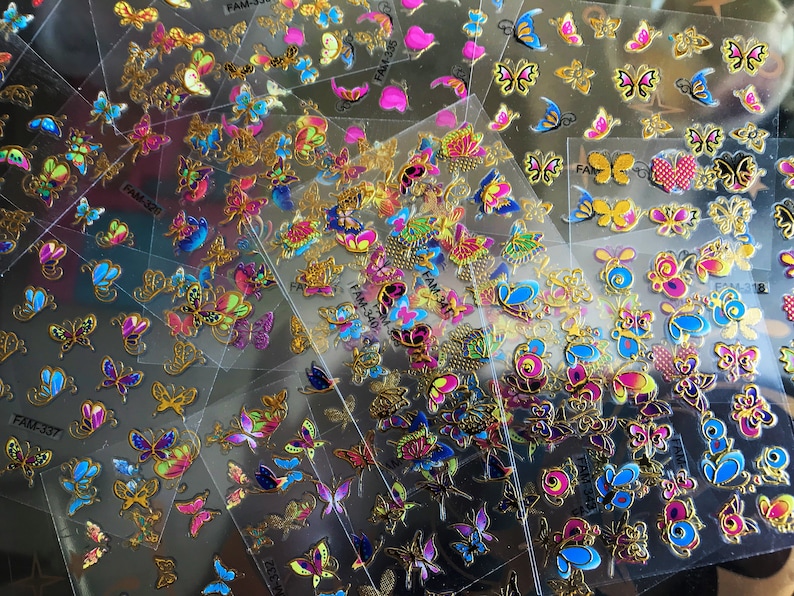 May include: Assortment of colorful butterfly nail stickers on clear plastic sheets. The stickers feature gold accents and various designs, including hearts, flowers, and swirls. The sheets are labeled with the product code "FAM-337".