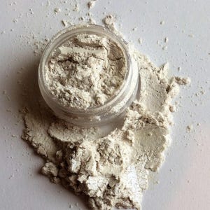 May include: A small clear jar filled with a white powder with a shimmery finish. The powder is spilling out of the jar onto a white surface.
