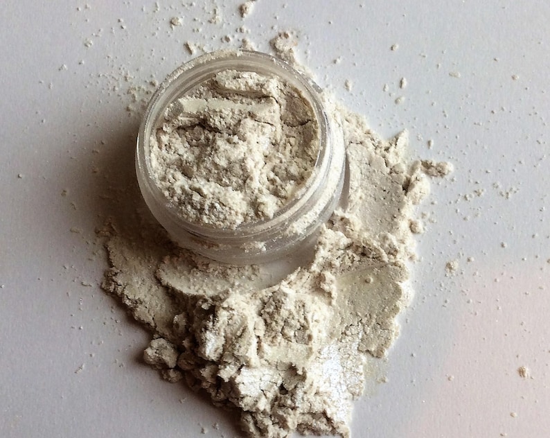 May include: A small clear jar filled with a white powder with a shimmery finish. The powder is spilling out of the jar onto a white surface.