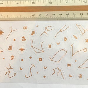 May include: A sheet of gold-colored constellation stickers with various zodiac signs, including Aries, Taurus, Gemini, Cancer, Leo, Virgo, Libra, Scorpio, Sagittarius, Capricorn, Aquarius, and Pisces. The stickers also include stars, moons, and other celestial symbols.