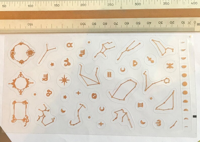 May include: A sheet of gold-colored constellation stickers with various zodiac signs, including Aries, Taurus, Gemini, Cancer, Leo, Virgo, Libra, Scorpio, Sagittarius, Capricorn, Aquarius, and Pisces. The stickers also include stars, moons, and other celestial symbols.