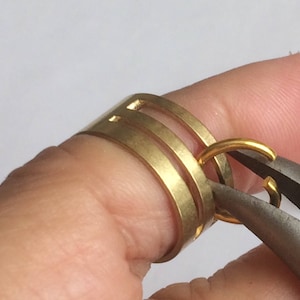 May include: Close-up of a gold-colored ring being worked on with pliers. The ring has a unique design with multiple bands and cut-out details. A smaller gold ring is being attached, showcasing jewelry craftsmanship.