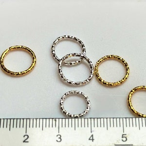 May include: Six jump rings, two gold and four silver, with a textured finish. The rings are arranged on a white surface with a ruler in the background.