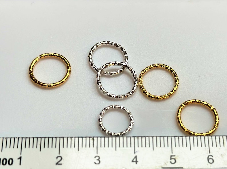 May include: Six jump rings, two gold and four silver, with a textured finish. The rings are arranged on a white surface with a ruler in the background.