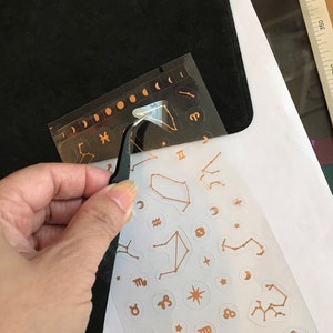 May include: A sheet of gold foil stickers with various zodiac signs and constellations. The stickers are on a white background and are being peeled off of a black sheet with a pair of tweezers.