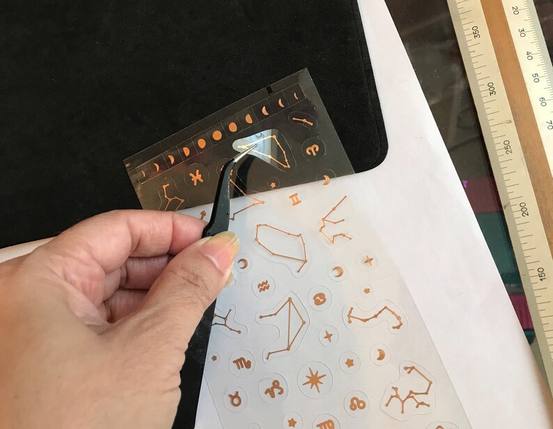 May include: A sheet of gold foil stickers with various zodiac signs and constellations. The stickers are on a white background and are being peeled off of a black sheet with a pair of tweezers.