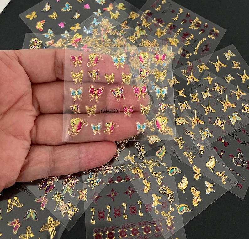 May include: Assortment of clear sheets with gold butterfly nail decals. Each sheet features a variety of butterfly designs with colorful accents. The sheets are labeled "FAM-320" and "FAM-521".