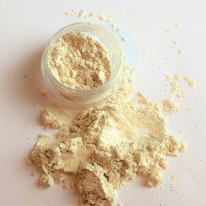 May include: A small clear jar filled with a light beige powder, with some of the powder spilled out onto a white surface.