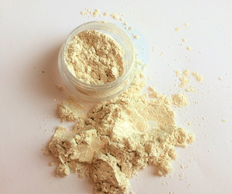 May include: A small clear jar filled with a light beige powder, with some of the powder spilled out onto a white surface.