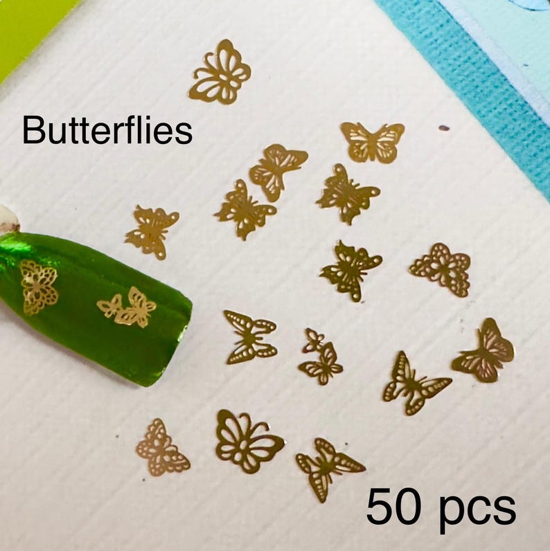 Op de afbeelding: Een set van 50 gouden vlinder nagel stickers. De stickers zijn gerangschikt op een wit oppervlak met een groene nagellak fles in de achtergrond. De tekst "Butterflies" en "50 pcs" is zichtbaar in de afbeelding.