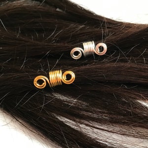 Op de afbeelding: Twee haarspiralen, een gouden en een zilveren, zijn te zien in een close-up afbeelding. De spiralen zijn om strengen donkerbruin haar gewikkeld.