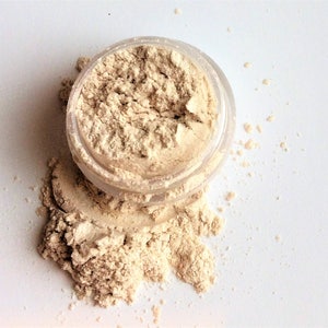 May include: A small clear jar filled with a light beige powder. The powder is spilling out of the jar onto a white surface.