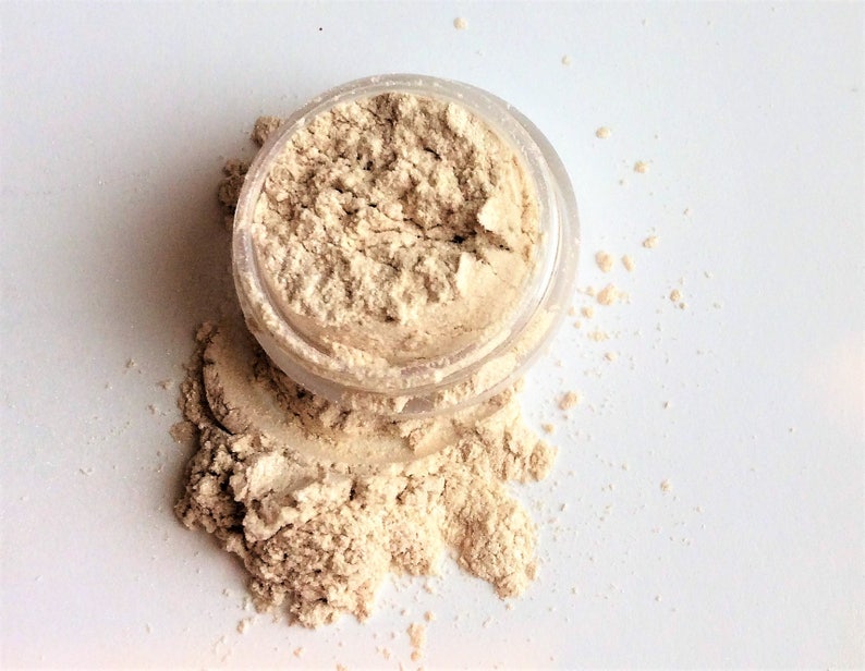 May include: A small clear jar filled with a light beige powder. The powder is spilling out of the jar onto a white surface.