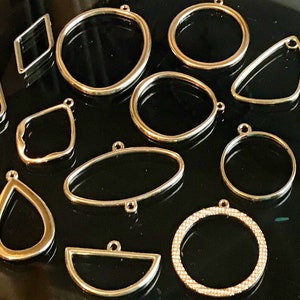 May include: Assortment of gold metal pendant frames in various shapes, including circles, ovals, diamonds, and teardrops. Some frames have a textured or beaded design.