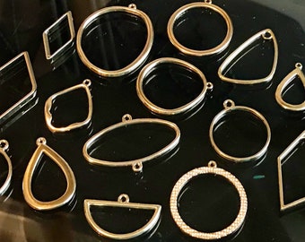 May include: A collection of gold-toned metal open-back pendant frames in various geometric shapes, including circles, ovals, diamonds, teardrops, and a semicircle. Each frame features a small loop at the top or side for attachment to jewelry.