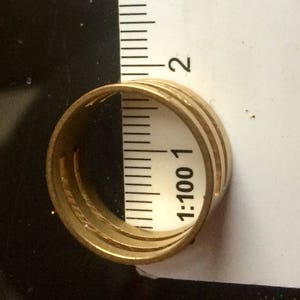 May include: A gold-colored ring with three bands is positioned next to a ruler. The ruler shows measurements in inches. The ring's design features a series of parallel lines, creating a textured appearance. The ring is set against a dark background.