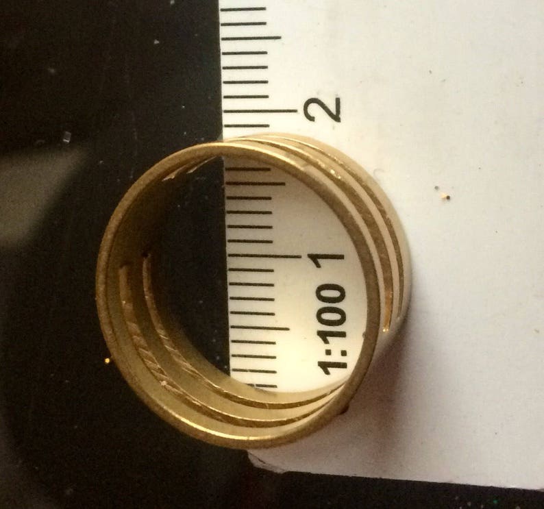 May include: A gold-colored ring with three bands is positioned next to a ruler. The ruler shows measurements in inches. The ring's design features a series of parallel lines, creating a textured appearance. The ring is set against a dark background.