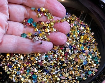 May include: A close-up of a hand holding a bowl filled with a variety of colorful rhinestones. The rhinestones are in various shapes and sizes, including round, square, and star-shaped. The rhinestones are a mix of gold, silver, red, blue, green, and clear.