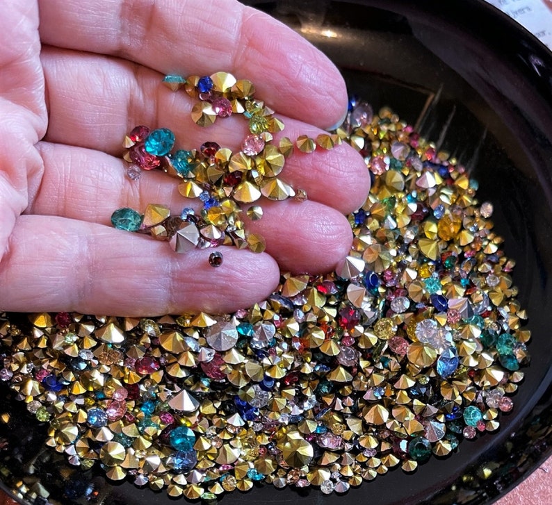 May include: A close-up of a hand holding a bowl filled with a variety of colorful rhinestones. The rhinestones are in various shapes and sizes, including round, square, and star-shaped. The rhinestones are a mix of gold, silver, red, blue, green, and clear.