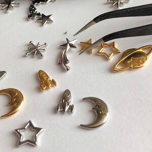 Może przedstawiać: Kolekcja srebrnych i złotych metalowych zawieszek z motywami kosmicznymi, w tym gwiazdami, planetami, księżycami i rakietami. Zawieszki są ułożone na białym tle z pęsetą.