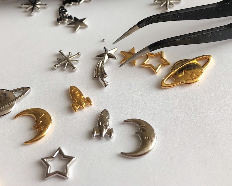 Może przedstawiać: Kolekcja srebrnych i złotych metalowych zawieszek z motywami kosmicznymi, w tym gwiazdami, planetami, księżycami i rakietami. Zawieszki są ułożone na białym tle z pęsetą.