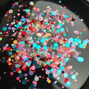 Pu&ograve; includere: Primo piano di una superficie nera ricoperta di glitter colorati. Il glitter &egrave; a forma di piccoli cerchi ed &egrave; un mix di rosso, rosa, blu, verde, oro e argento.