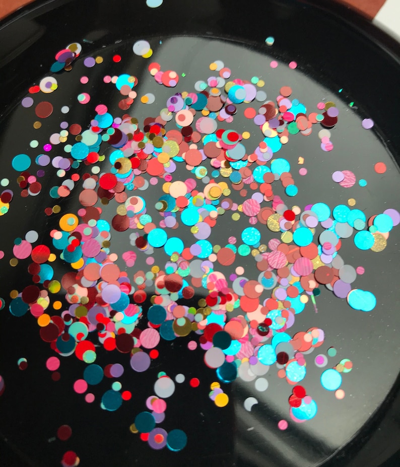 Pu&ograve; includere: Primo piano di una superficie nera ricoperta di glitter colorati. Il glitter &egrave; a forma di piccoli cerchi ed &egrave; un mix di rosso, rosa, blu, verde, oro e argento.