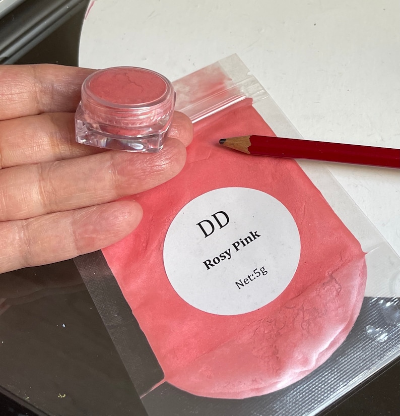 May include: A small clear jar filled with a rosy pink powder. The jar is labeled "DD Rosy Pink Net: 5g".