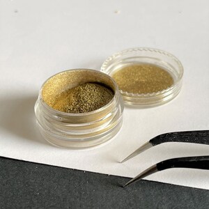 K&ouml;nnte beinhalten: Zwei kleine, transparente Plastikgl&auml;ser mit goldfarbenem Pulver stehen auf einer wei&szlig;en Oberfl&auml;che. Ein Glas ist ge&ouml;ffnet und gibt den Blick auf das Pulver frei. Eine schwarze Pinzette liegt daneben, was auf die Verwendung in einem Bastel- oder Sch&ouml;nheitsprojekt hindeutet.