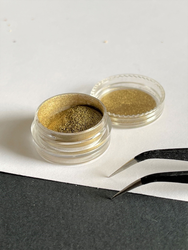 K&ouml;nnte beinhalten: Zwei kleine, transparente Plastikgl&auml;ser mit goldfarbenem Pulver stehen auf einer wei&szlig;en Oberfl&auml;che. Ein Glas ist ge&ouml;ffnet und gibt den Blick auf das Pulver frei. Eine schwarze Pinzette liegt daneben, was auf die Verwendung in einem Bastel- oder Sch&ouml;nheitsprojekt hindeutet.