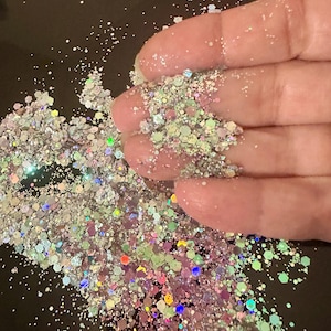 May include: Close-up of iridescent glitter in various shapes and sizes. The glitter is a mix of silver, pink, green, and blue, creating a shimmering effect. The glitter is held in a hand, with some particles scattered around.