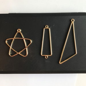 Op de afbeelding: Drie gouden metalen frames in de vorm van een ster, een rechthoek en een driehoek. De frames bevinden zich op een zwart oppervlak.