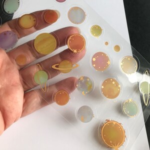 Op de afbeelding: Een vel met transparante stickers met gouden contouren met verschillende planeten, sterren en manen. De stickers zijn perfect om een ​​hemelse touch toe te voegen aan planners, dagboeken en andere knutselprojecten.
