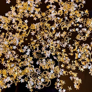 Op de afbeelding: Een verzameling gouden en zilveren metalen bedels in de vorm van sterren, manen en geometrische vormen. De bedels zijn verspreid over een zwart oppervlak.