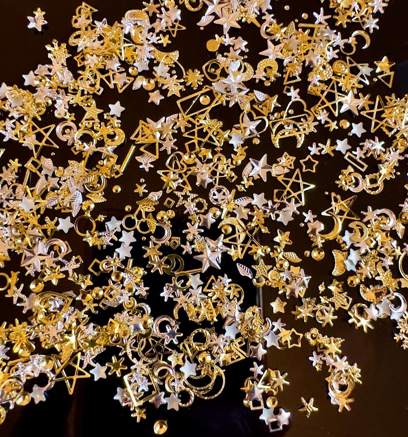 Op de afbeelding: Een verzameling gouden en zilveren metalen bedels in de vorm van sterren, manen en geometrische vormen. De bedels zijn verspreid over een zwart oppervlak.