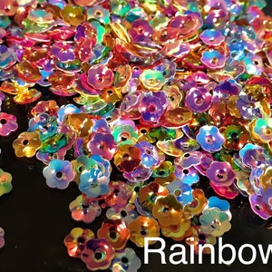 Può includere: Un grande mucchio di paillettes iridescenti a forma di fiore arcobaleno. Le paillettes sono sparse su una superficie nera. La parola "Rainbow" è scritta in lettere bianche nella parte inferiore dell'immagine.