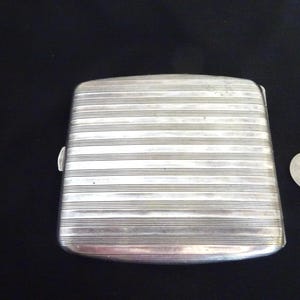 May include: A silver cigarette case with a ribbed design. The case is closed and has a smooth, polished finish.