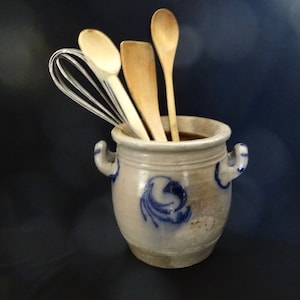 May include: A stoneware pot with blue floral patterns contains a whisk, a spatula, and two wooden spoons. The pot has two small handles and is set against a dark blue backdrop. Kitchen utensils.