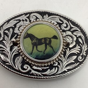 May include: Oval silver-tone belt buckle with a black horse illustration in the center. The buckle has a detailed floral and scroll design with black enamel accents. The horse is depicted in a running pose against a green and blue background.