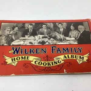 May include: Vintage red cookbook titled "The Wilken Family Home Cooking Album." The cover features a black and white photo of a family gathered around a dining table. The title is in a decorative font.